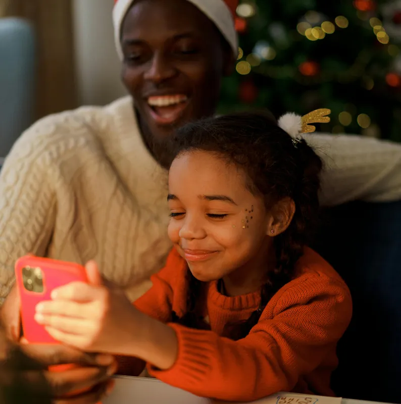 Vater mit Weihnachtsmütze und Tochter schauen lachend auf ein Handy.